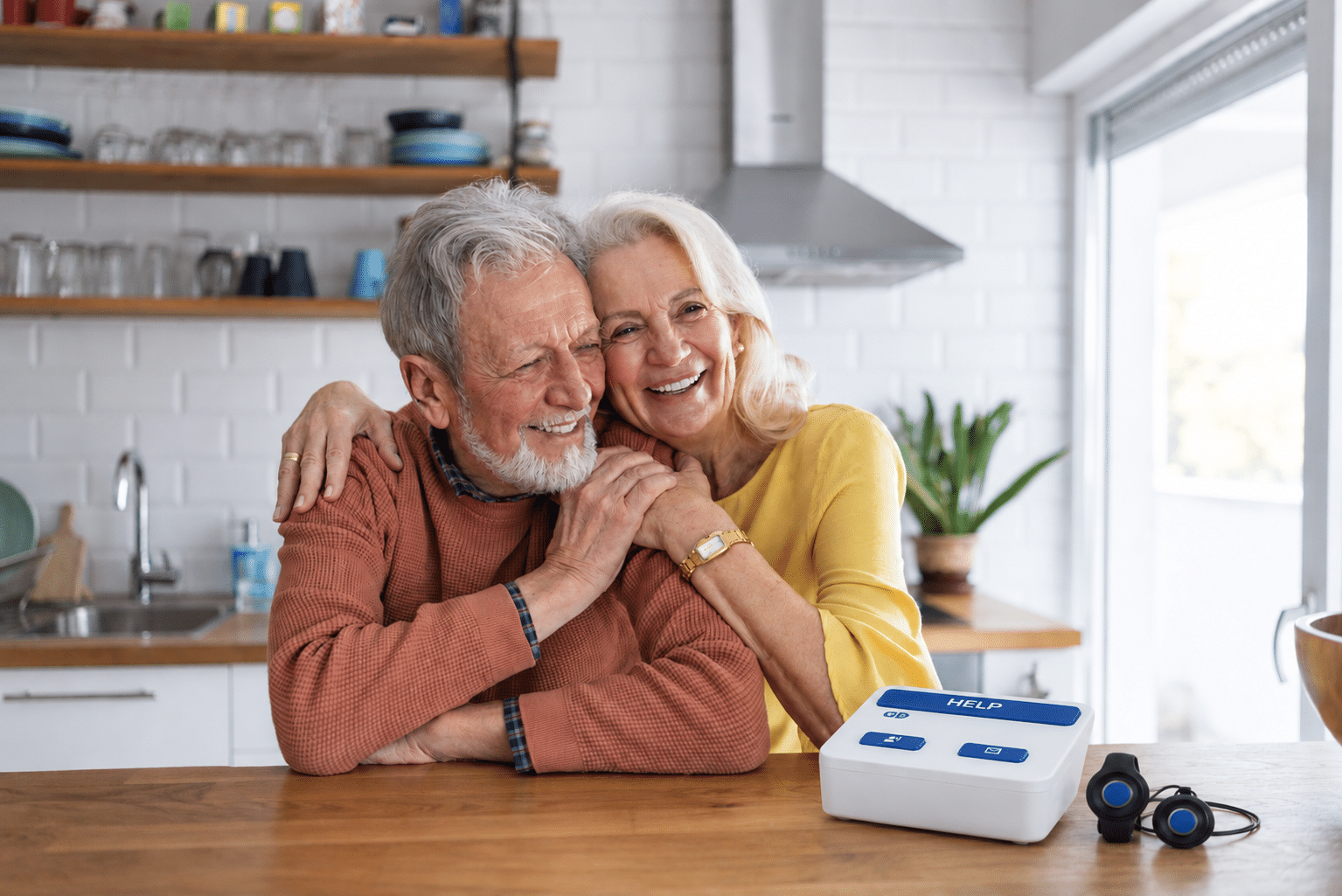 Smiling senior couple sitting together at home with a medical alert base unit on the table, illustrating a medical alert without a landline.